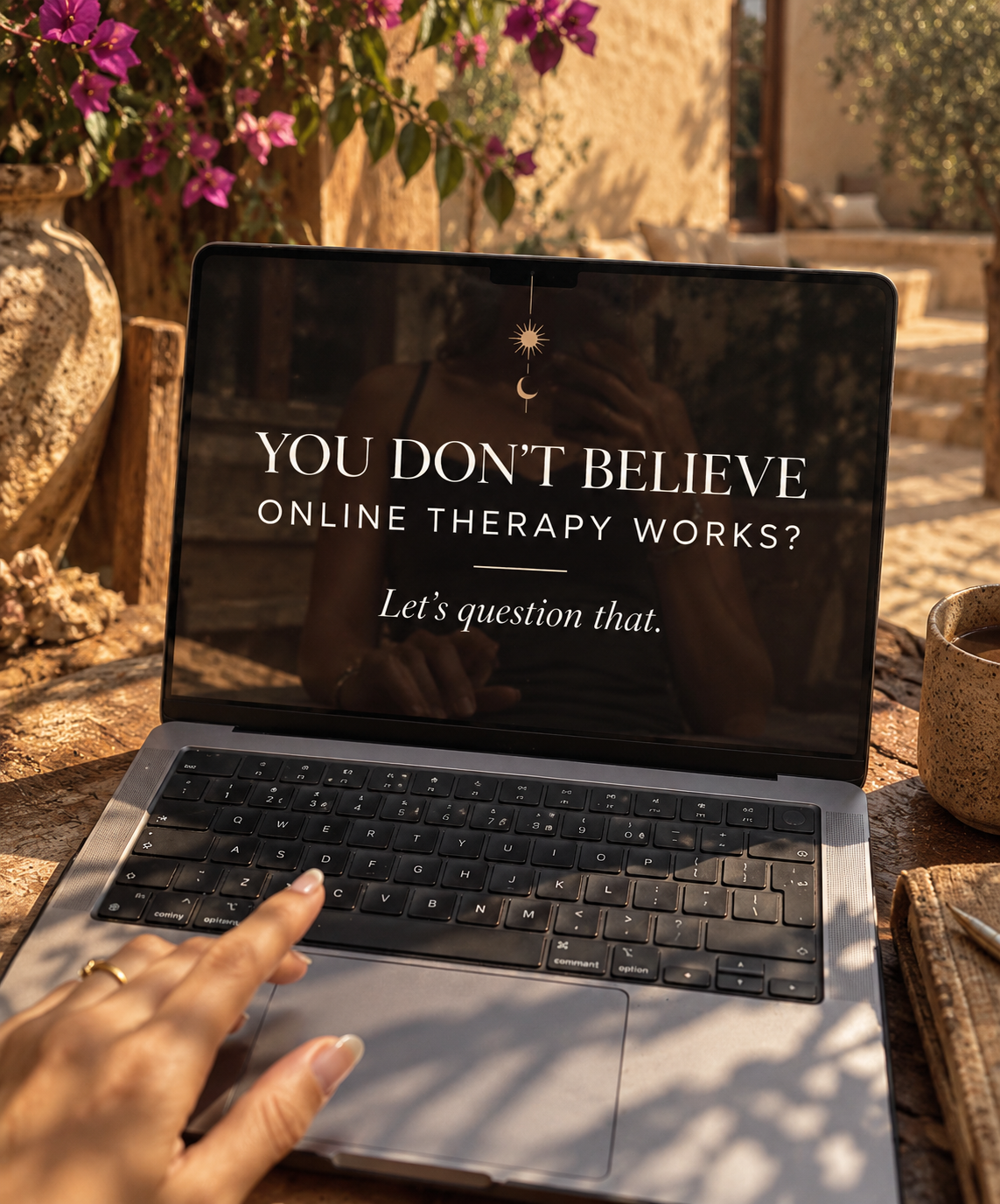 A laptop open on a rustic wooden table in a sunlit courtyard, its screen reading: You don't believe online therapy works? Let's question that. A hand rests on the trackpad, beside a textured ceramic mug, a notebook with a pen, and a clay urn of pink bougainvillea.