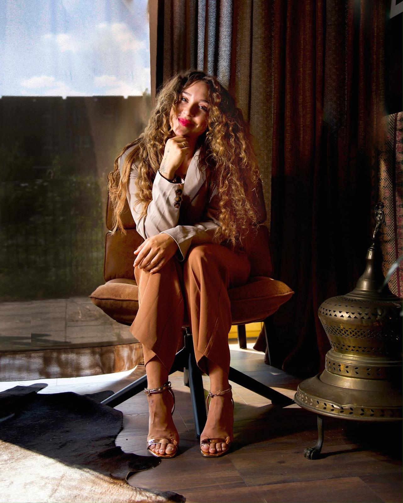 Marije Andrea seated in a brown swivel chair beside a brass lantern, daylight from a tall window behind her